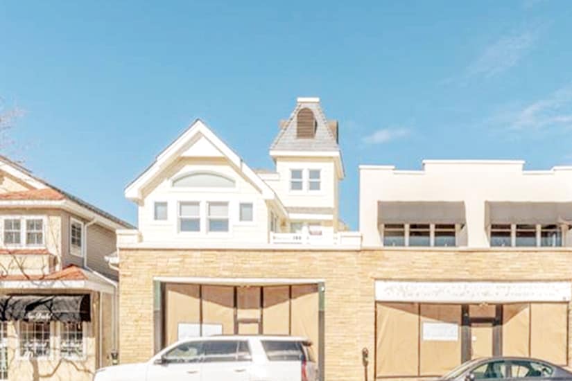 A contemporary building is displayed with a mix of architectural styles, showcasing a peaked roof and large windows. The facade features light-colored stone and a modern entrance, while a parked vehicle is visible in front, contributing to the vibrant neighborhood setting.