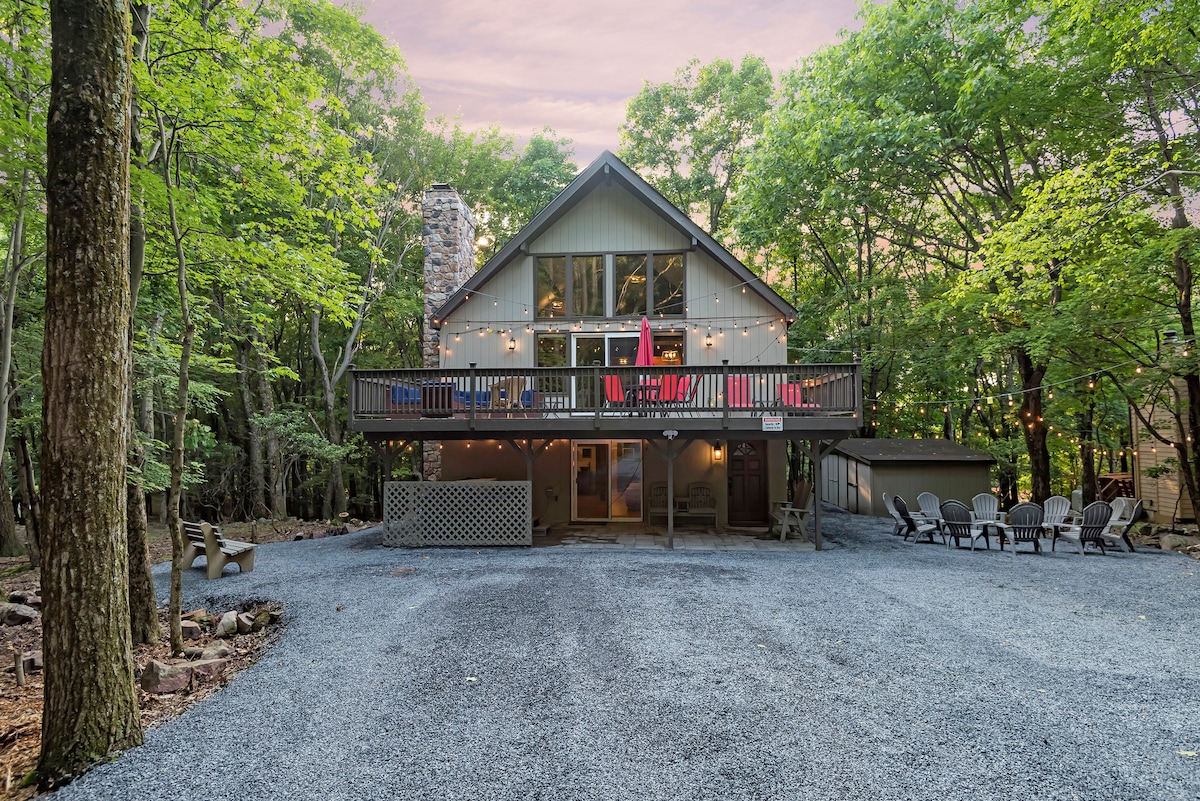 The cabin is nestled among lush trees, featuring a spacious deck adorned with lights and seating. A stone chimney rises from the side, while gravel leads up to the entrance. Outdoor chairs are positioned in front, inviting relaxation in a serene environment.
