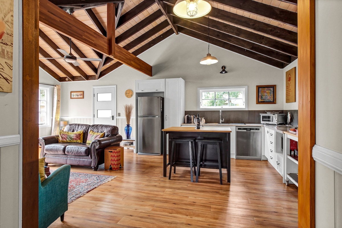 An open-concept living area features a comfortable leather sofa and a wooden dining table surrounded by stylish black stools. Natural light floods in through large windows, highlighting the high, exposed beam ceiling and wooden flooring. A well-equipped kitchen with modern appliances is visible in the background.