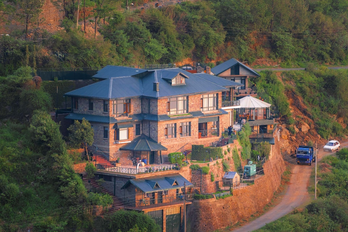 The expansive property is set amidst lush greenery on a hillside, showcasing a multi-level structure with a blend of stone and modern design. A cozy glass-walled sunroom can be seen, along with various outdoor seating areas and pathways leading to the entrance.