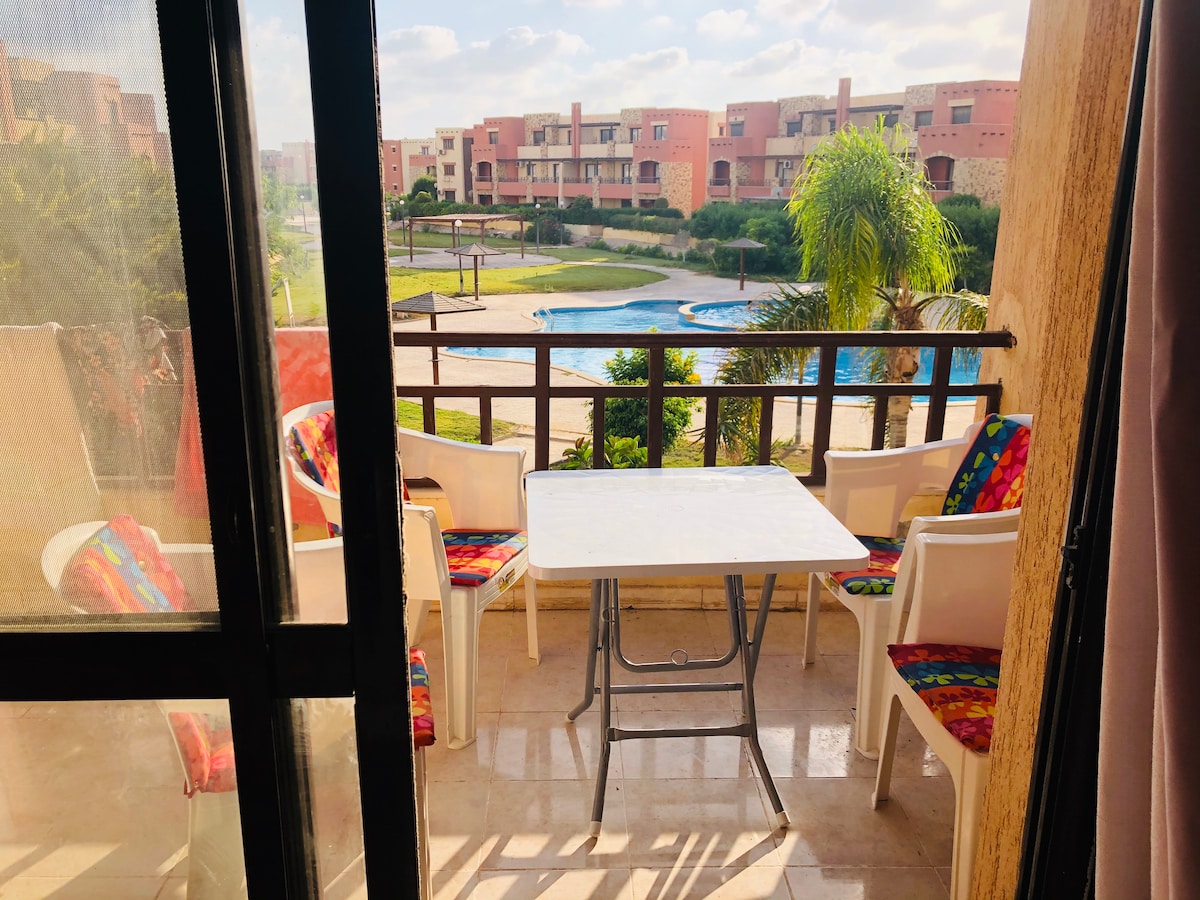 A private balcony is showcased, featuring a table and several chairs dressed in colorful cushions. The view overlooks a serene pool area surrounded by lush greenery, with additional buildings visible in the background under a clear sky.