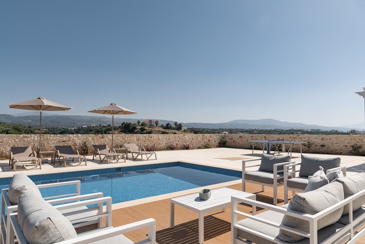 An inviting outdoor living space features a private swimming pool surrounded by sun loungers and umbrellas. Comfortable seating areas with gray cushions are arranged on a wooden deck, offering scenic views of the lush landscape and distant mountains under a clear blue sky.