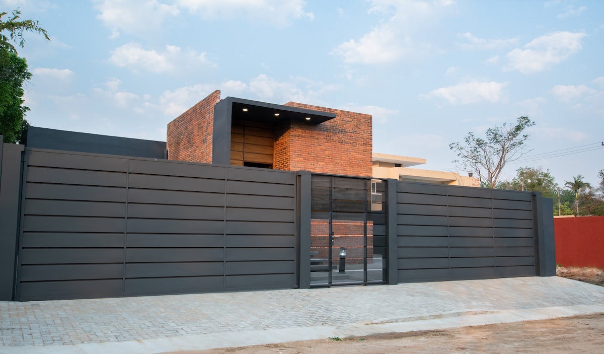 The exterior showcases a modern brick house with a sleek gray fence, presenting a minimalistic yet welcoming front. The design is complemented by a clear pathway leading to the entrance, surrounded by a well-maintained landscape.