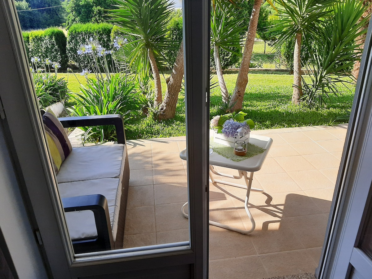 A charming outdoor terrace is visible through sliding glass doors, featuring a small table with a drink and a cloth, accompanied by a comfortable sofa. Lush greenery surrounds the space, including palm trees and flowering plants, creating a serene atmosphere.