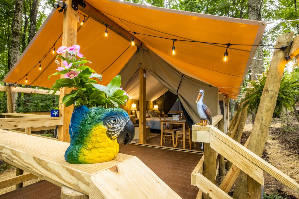 A welcoming glamping tent is set against a backdrop of trees, featuring an orange canopy and warm lighting. A vibrant planter shaped like a parrot holds flowers at the entrance, while a decorative bird figurine is positioned nearby, enhancing the natural surroundings.
