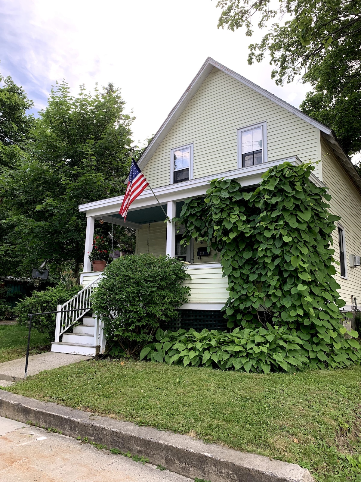 A charming two-story house features a welcoming front porch with steps leading to the entrance. Lush green vines decorate the exterior, while an American flag adds a touch of patriotism. The surrounding yard is neatly maintained with grass and landscaping.