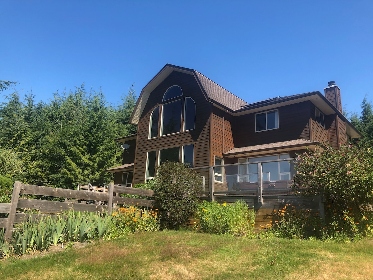 A two-story wooden house is set against a clear blue sky, surrounded by lush greenery and blooming flowers. Large windows allow for plenty of natural light, while a wooden fence encloses the garden area. The structure blends harmoniously with the natural surroundings.