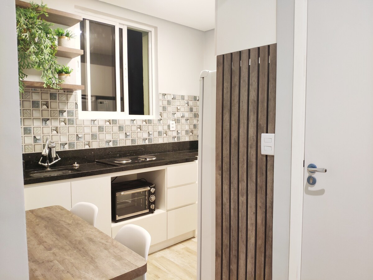 A well-equipped kitchenette is visible, featuring a blend of light cabinetry and a patterned tile backsplash. A small dining table with white chairs is positioned nearby. Natural light filters through a window above the sink, creating a bright and inviting space.