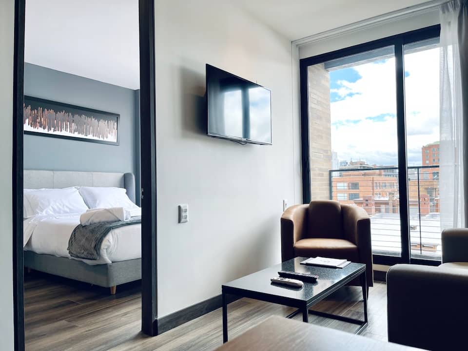 A modern living area features a comfortable seating arrangement with two chairs and a coffee table. A spacious bed is visible through an open doorway, complemented by a wall-mounted television. Floor-to-ceiling windows allow natural light to illuminate the space.