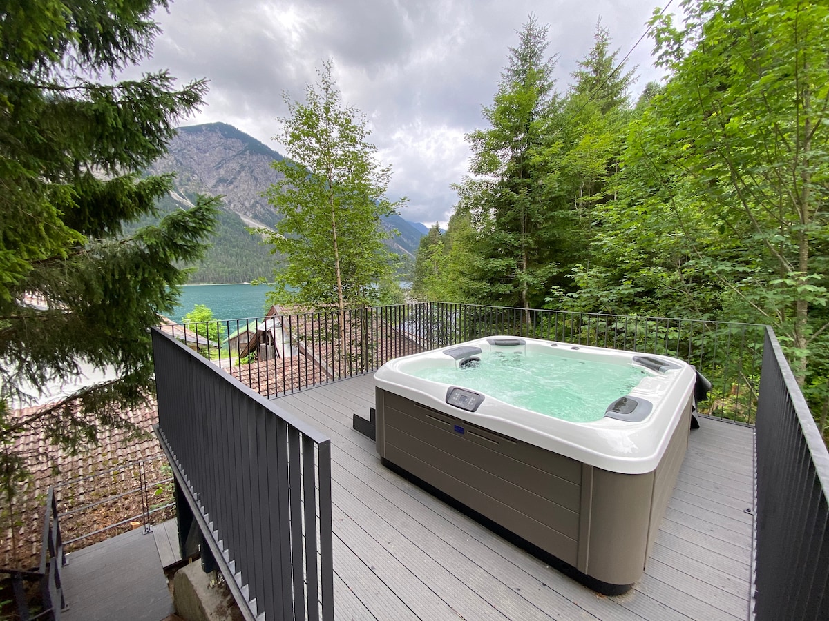 A spacious hot tub is situated on a wooden terrace surrounded by trees, offering a view of the nearby lake and mountains. The serene environment creates an inviting setting for relaxation, with the hot tub's water shimmering under the overcast sky.