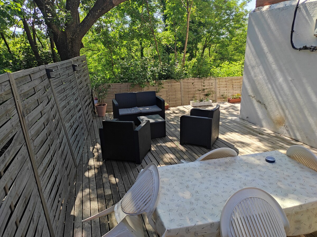 An outdoor terrace is showcased, featuring a wooden deck and surrounded by greenery. A seating area with black wicker chairs and a coffee table is present, along with a dining table. Sunlight filters through the trees, creating a serene setting.