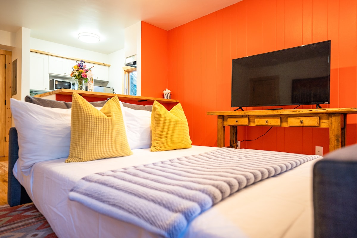 A spacious living area features a queen-sized sleeper sofa adorned with two yellow accent pillows. A gray throw blanket is neatly placed at the foot of the sofa. The room is complemented by a striking orange wall and a large Smart TV positioned on a wooden table.