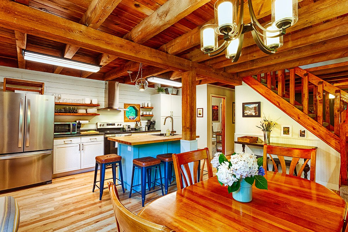 An open concept kitchen and dining area feature natural wood beams and a warm tone. A large dining table is centered in the foreground, surrounded by wooden chairs. The kitchen showcases modern appliances, with a central island offering additional seating and space for meal preparation.