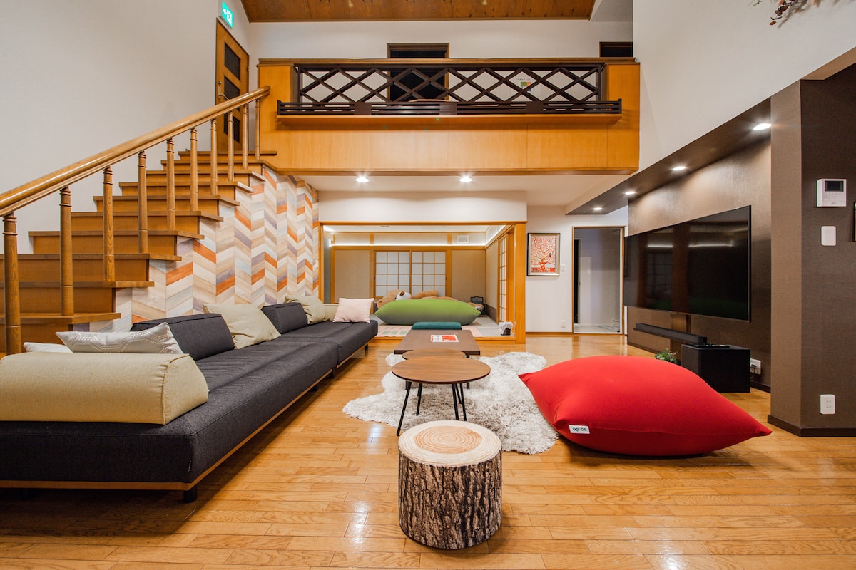 A spacious living area features a modern, low-profile sofa with light-colored cushions. A coffee table sits in front, with two large, colorful floor cushions nearby. The hardwood floor and ascending staircase with wooden railings complement the warm tones of the wall decorations and lighting.