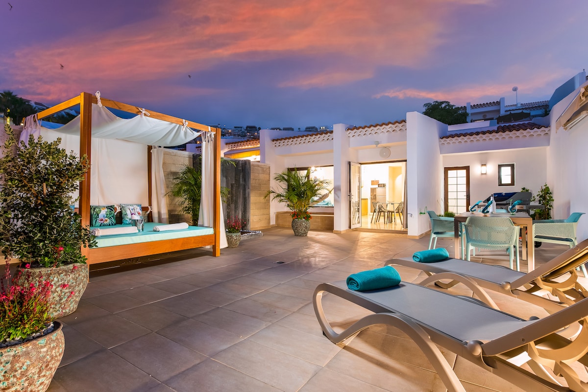 An expansive chillout terrace features a wooden Balinese bed with soft cushions, surrounded by decorative plants. Sun loungers are positioned nearby, and a dining area is visible in the background, illuminated by the warm hues of a sunset sky.