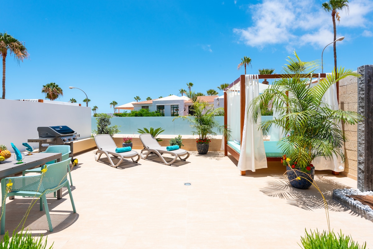 An expansive terrace features comfortable loungers and a barbecue area, surrounded by tropical plants. A water fountain adds a serene touch, while sheer curtains create a private ambiance. The tranquil outdoor space is bathed in sunlight, showcasing a perfect spot for relaxation and entertainment.