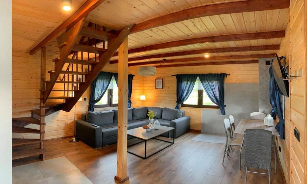 A welcoming living area features a comfortable sectional sofa facing a coffee table, with ample natural light from windows adorned with blue curtains. A dining table with chairs is positioned nearby, while a wooden staircase leads to the upper level. Rustic wooden walls create a cozy ambiance.