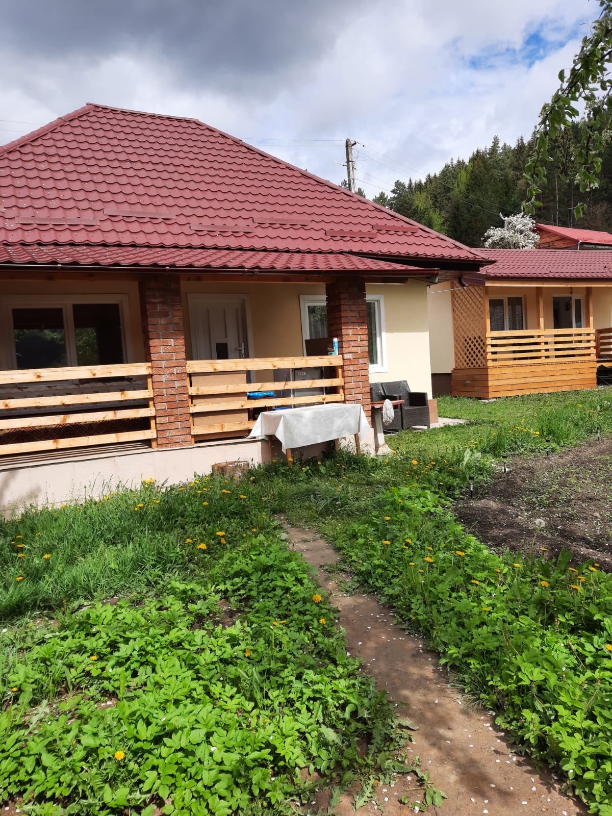 Two cottages are situated side by side, each featuring a covered porch with wooden railings. A narrow path of dirt and greenery leads between the cottages, surrounded by patches of grass and wildflowers. The tree line and cloudy sky are visible in the background.
