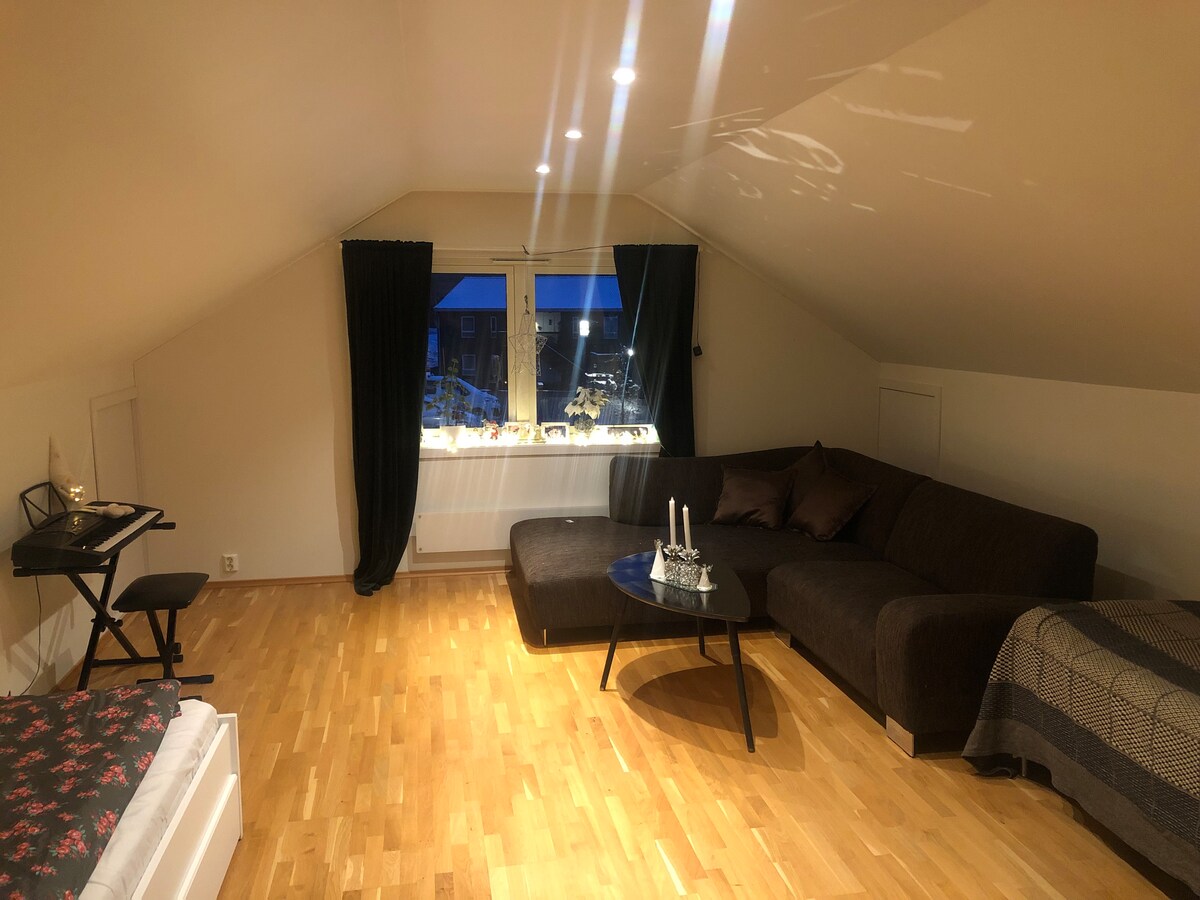 A spacious third-floor room features a large, comfortable sofa and a small table. A cozy bed with a patterned bedspread occupies one corner, while natural light filters through the front-facing window adorned with dark curtains. A compact writing desk is also visible.