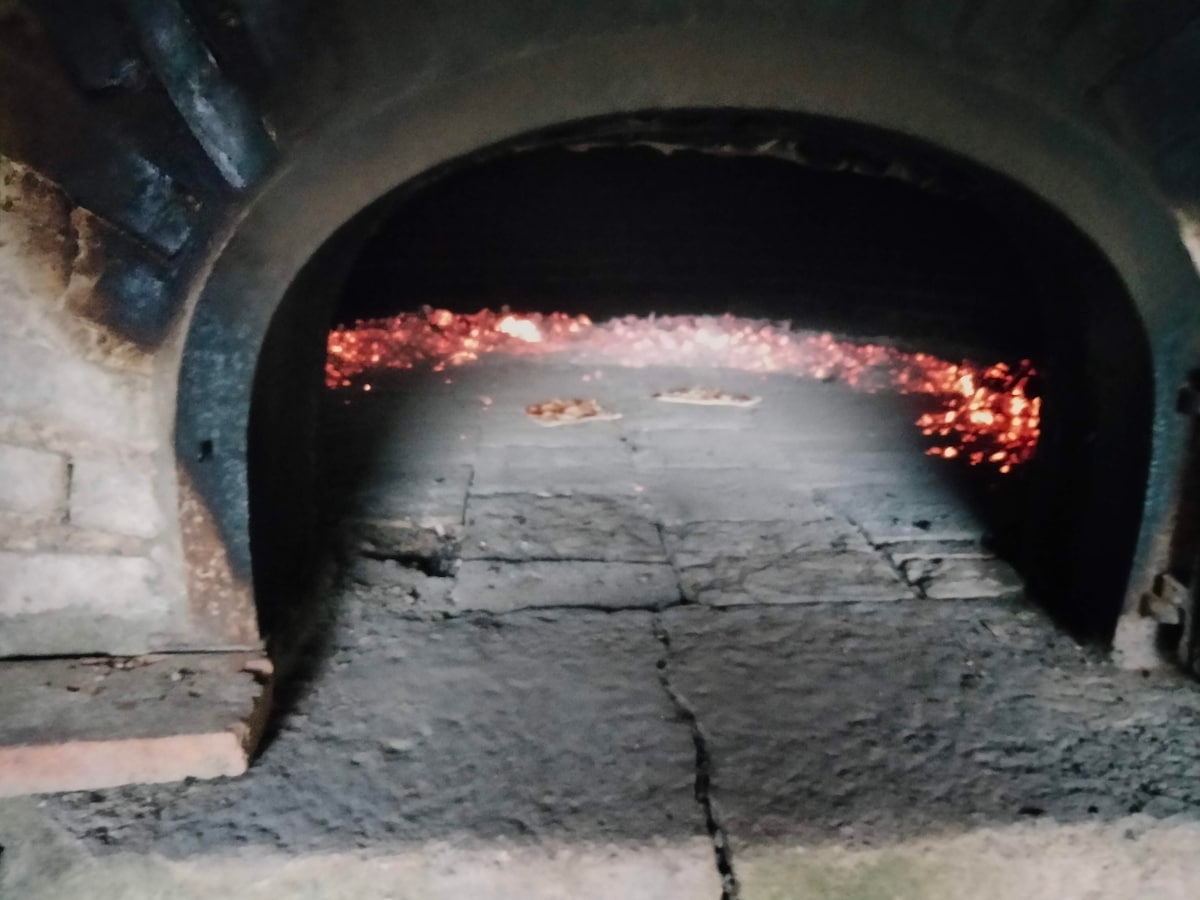 An old stone bakery oven is presented, showcasing a darkened interior with glowing embers visible at the back. The arched entrance is framed by aged bricks, highlighting the historical character of the structure. The stone floor remains in view, displaying a timeworn texture.