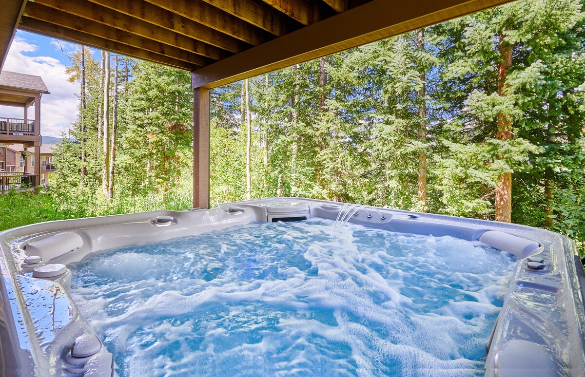 A spacious hot tub is situated under a covered area, offering a relaxing environment surrounded by lush greenery. The bubbling water invites guests to unwind while enjoying the natural surroundings.