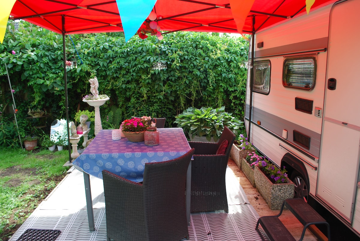 A shaded outdoor seating area features a table draped with a blue tablecloth, surrounded by black wicker chairs. Colorful bunting adds a festive touch overhead. Lush greenery and flowering plants are visible in planters, enhancing the serene atmosphere next to the caravan.