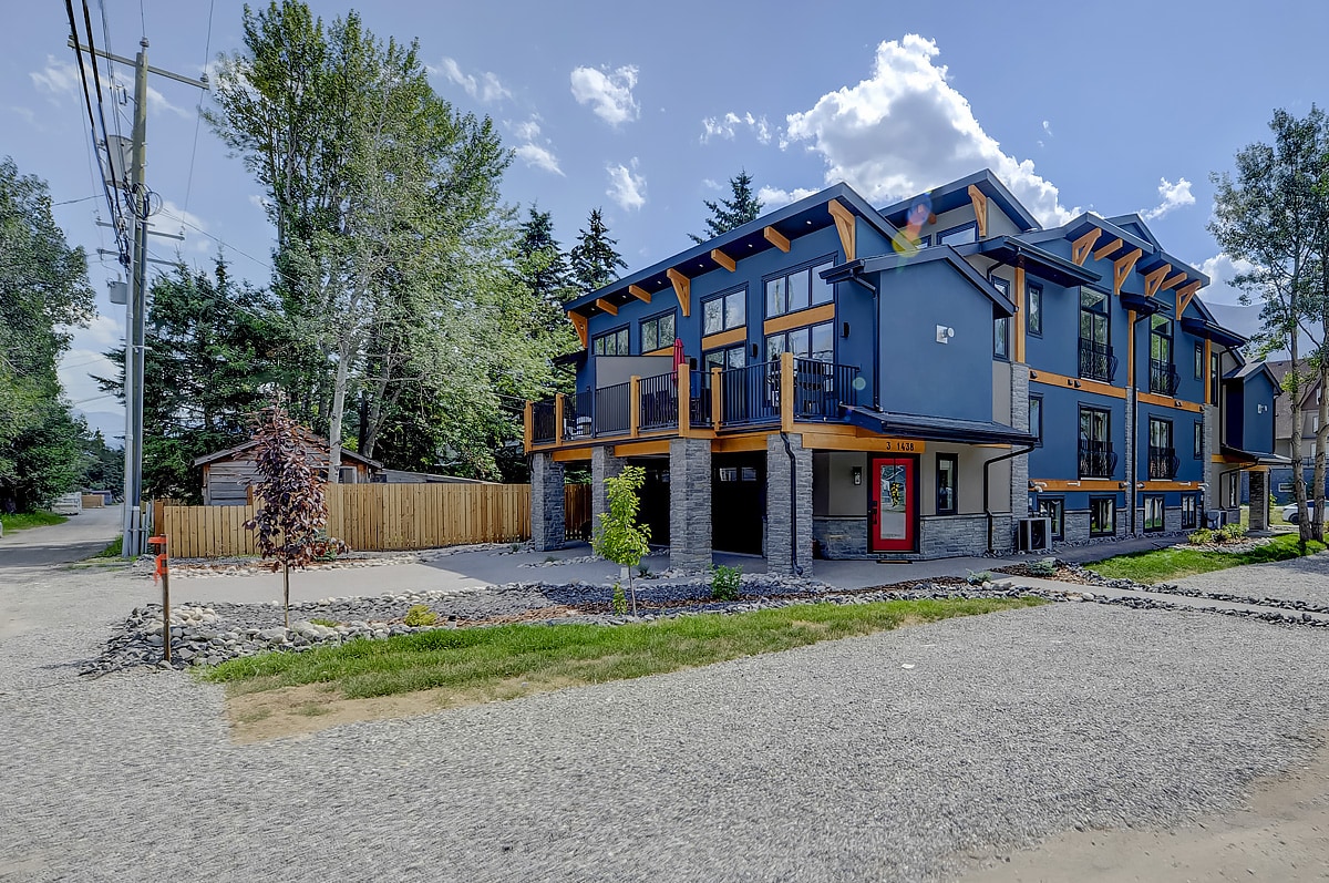 The exterior view showcases a newly built multi-unit home with a modern design. Colorful accents are presented against a blue facade, and inviting balconies are visible on multiple levels. A gravel path leads to the entrance, framed by green landscaping and nearby trees.