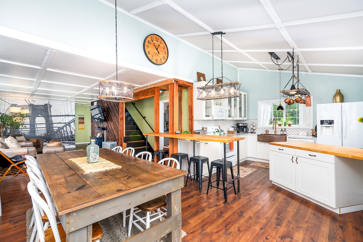 An open-concept kitchen and dining area features a large wooden table surrounded by white chairs, complemented by dark barstools. A clock hangs on the wall above, and light fixtures provide illumination. The space leads to a well-equipped kitchen, enhancing the communal atmosphere.