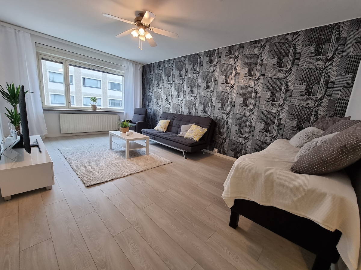A spacious living area features a dark patterned accent wall and large windows, allowing natural light to filter in. A light-colored area rug lies on the floor, complementing a low coffee table surrounded by a sofa and additional seating. Decorative plants add a touch of greenery.