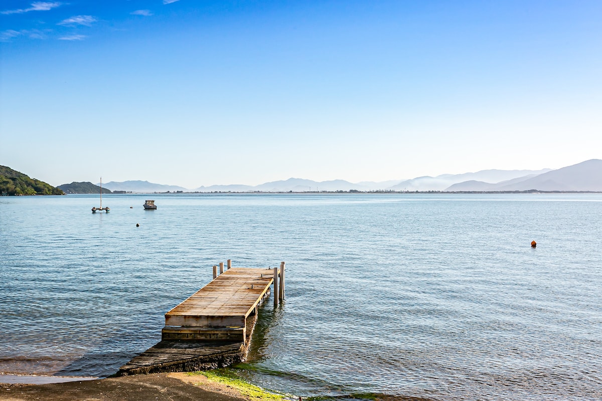 A wooden dock extends over calm water, leading towards distant mountains under a clear blue sky. The peaceful scene captures small boats anchored nearby, with gentle ripples reflecting the surrounding landscape.