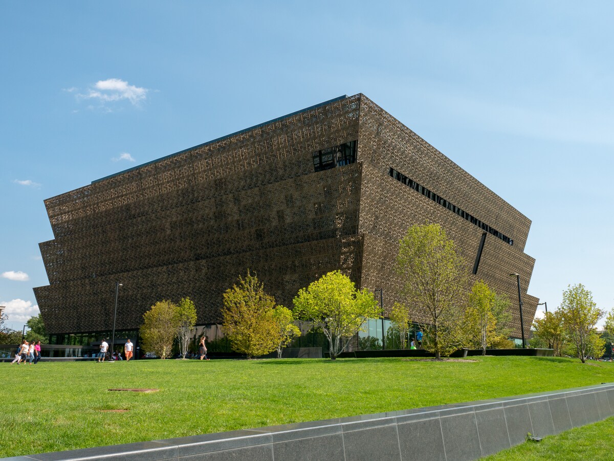 A modern architectural structure showcases a unique geometric design with a textured exterior. Surrounding the building, manicured green lawns and trees provide natural beauty. Visitors can be seen strolling along the pathways, enjoying the space on a clear, sunny day.