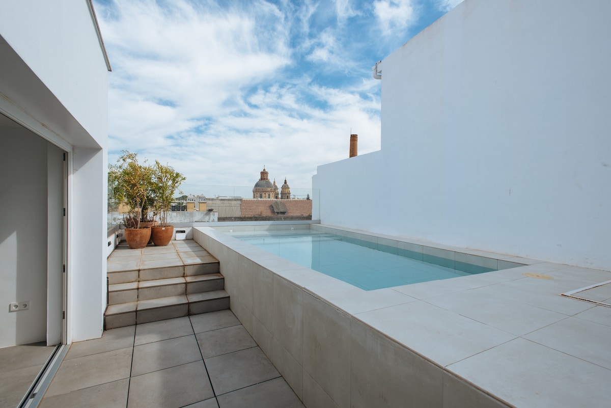 A serene rooftop terrace features a small swimming pool surrounded by light-colored tiles. Steps lead up to the pool area, with a few planters visible. The view captures nearby rooftops and church domes under a partly cloudy sky.