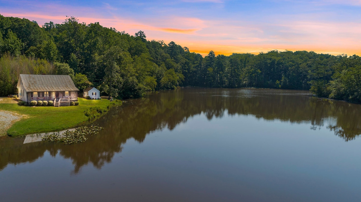 A serene view showcases a lake surrounded by lush greenery, with a modest home seated on the shore. The calm water reflects the surrounding trees and the evening sky, creating a peaceful atmosphere. A small shed is visible near the water's edge.