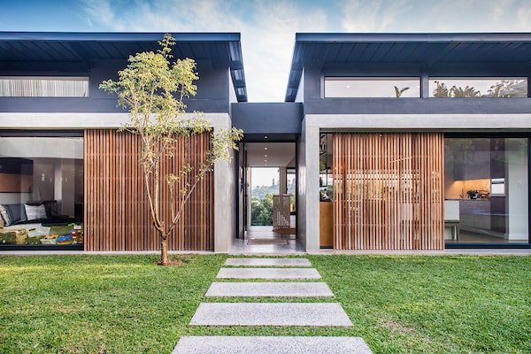 The entrance to the family home features a pathway of large stone slabs leading through a grassy area. Dual modern structures with wooden slatted facades flank the pathway, offering a glimpse into the interior spaces designed for open living and seamless connections with the outdoors.