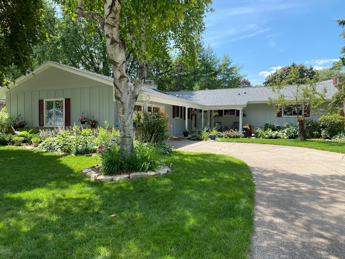 A spacious ranch-style home is surrounded by well-maintained gardens and greenery. The entrance features a wide driveway, leading to a covered porch with flower planters. Sunlight brightens the exterior walls while the trees provide shaded areas in the yard.