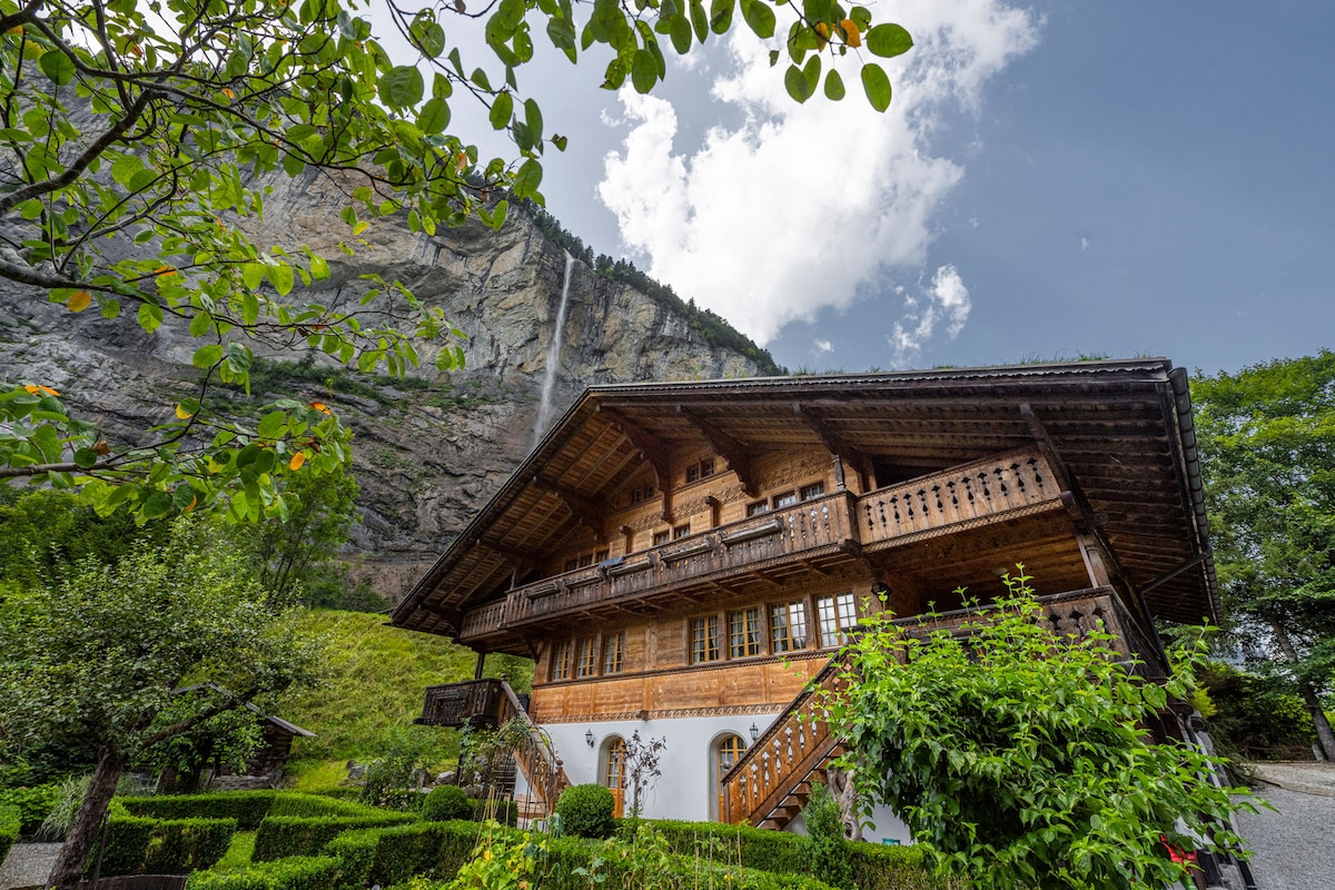 The wooden chalet features intricate balcony designs and large windows, set against a backdrop of a towering mountain and waterfall. Lush greenery surrounds the property, enhancing the natural beauty of the setting. The structure showcases a blend of traditional architecture and serene landscape.