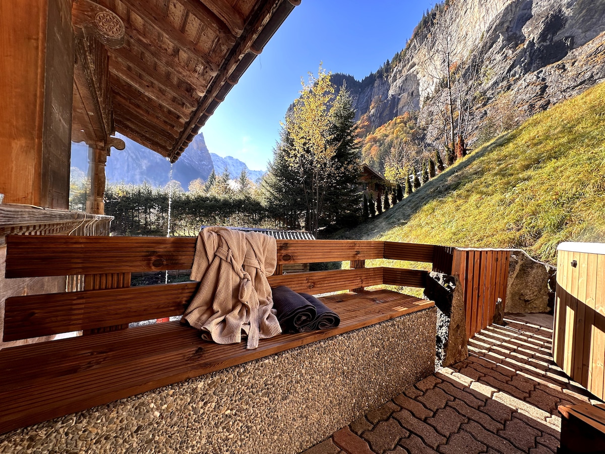 A wooden bench is positioned on a stone patio, providing a comfortable spot to relax. Soft towels are neatly folded on the bench. In the background, a lush green hillside rises towards rocky mountains, under a clear blue sky.