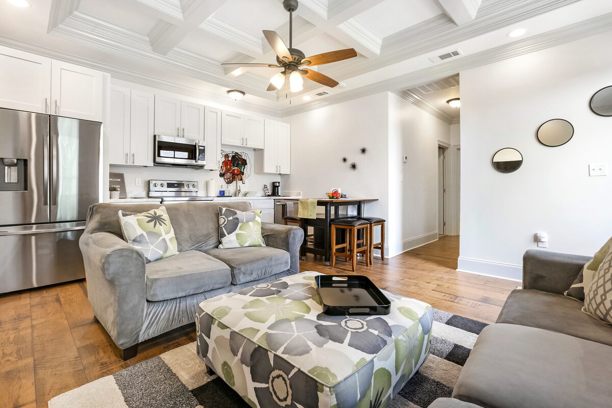 A light-filled living area features a coffered ceiling and a comfortable gray sofa adorned with decorative pillows. A stylish ottoman with floral patterns adds to the decor. The open kitchen showcases modern appliances, including a stainless-steel refrigerator and a dining table in the background.