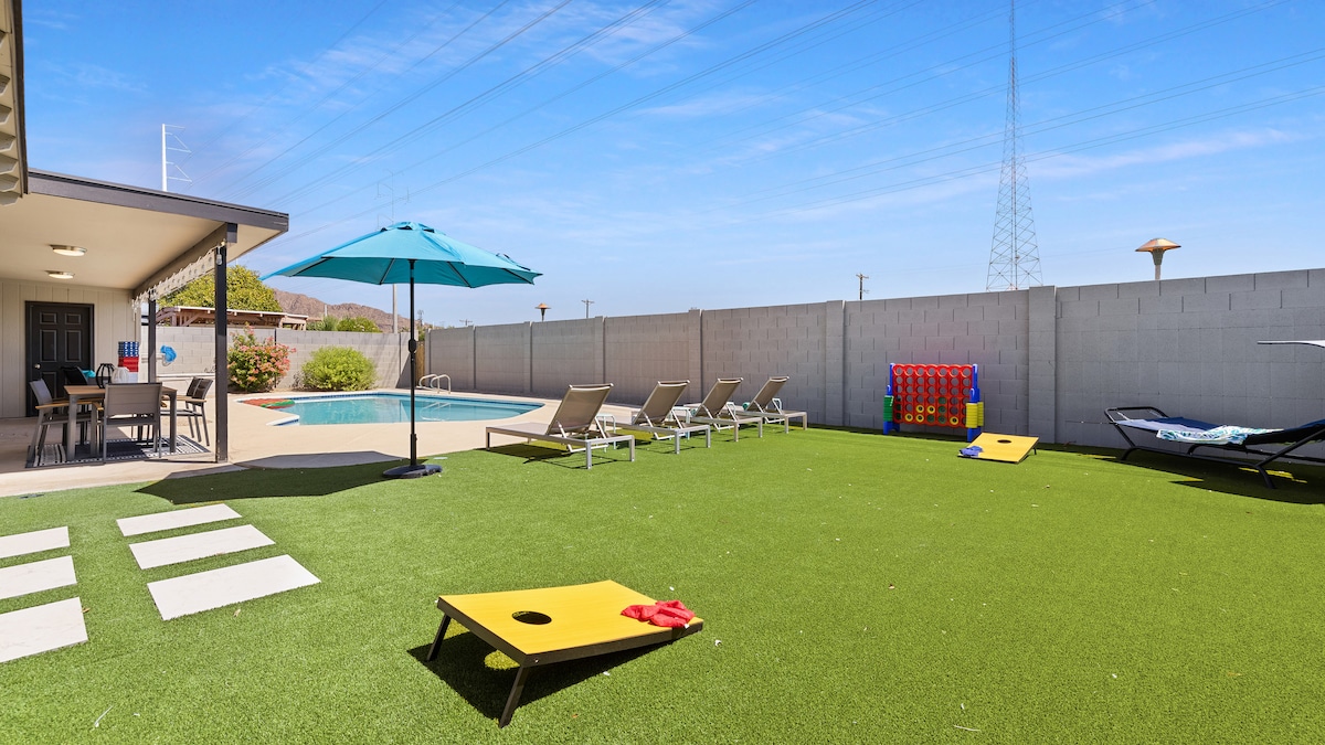 A spacious backyard area features a heated pool surrounded by lounge chairs and shaded by a large umbrella. Colorful yard games are available on the artificial turf, with a covered patio offering dining and seating options under a clear blue sky.