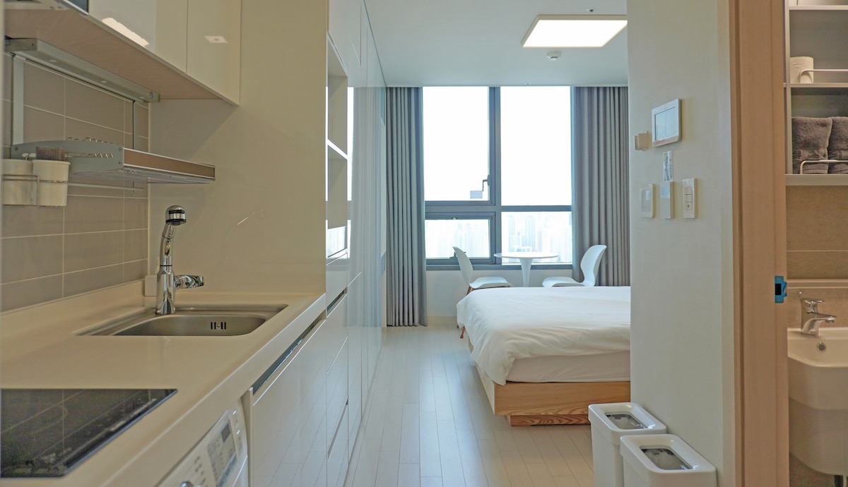 The image showcases a modern, open layout, featuring a kitchenette on the left with a sleek sink and stovetop. A spacious bed is positioned in the background, accompanied by large windows allowing natural light. Two light-colored chairs are visible near the window, creating a cozy seating area.