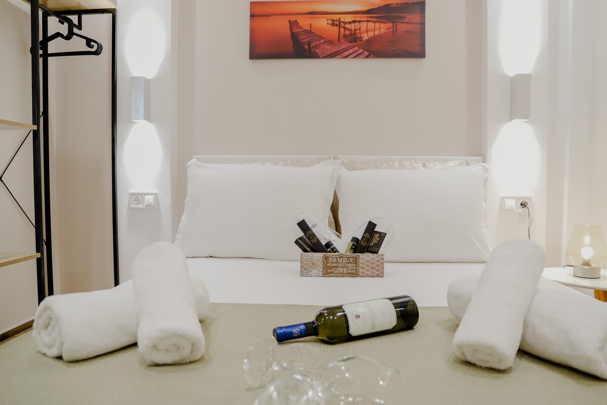 A spacious bed with crisp white linens is set against a warm-toned wall art piece depicting a scenic seascape. A wooden crate holds bottles of wine, while neatly rolled towels are placed on either side. Soft lighting from wall fixtures adds a gentle glow to the room.