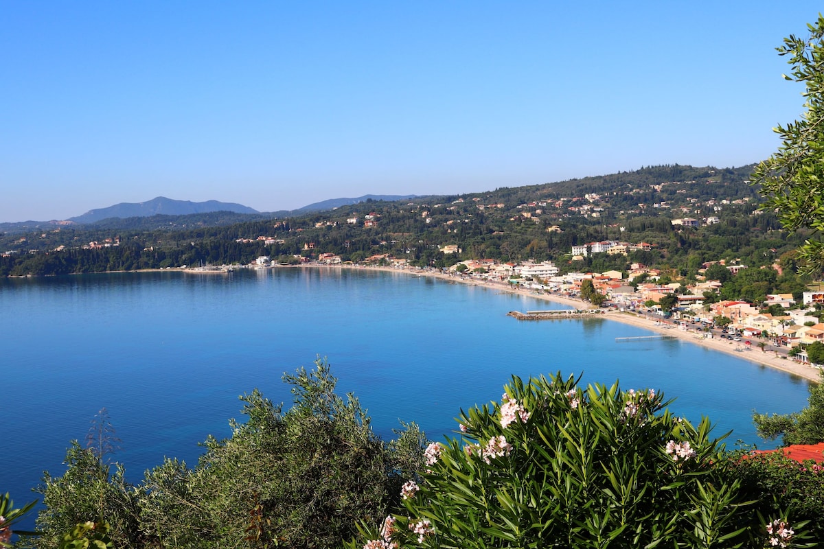 A panoramic view of a tranquil bay showcasing clear blue waters and a sandy beach, bordered by lush greenery. The coastal settlement features a variety of buildings and houses, nestled against a backdrop of rolling hills under a clear blue sky.