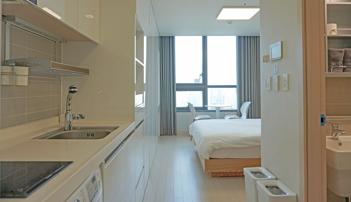 A well-lit room showcasing a spacious bed with crisp linens. A kitchenette featuring modern appliances is visible, along with a sleek countertop. Two chairs are positioned near large windows, allowing natural light to fill the space. The overall design emphasizes simplicity and functionality.