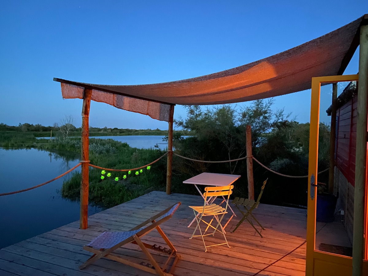 A covered terrace is visible, featuring a small table and two folding chairs, set against the backdrop of a tranquil body of water. Soft lighting adds warmth to the scene, with greenery surrounding the area, creating a calm natural setting.