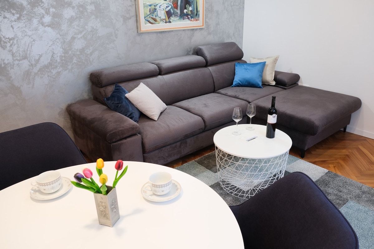 A comfortable seating area features a dark grey sectional sofa adorned with various cushions in different shades. A round white coffee table is placed centrally, accompanied by two black upholstered chairs. A small vase with colorful tulips adds a subtle touch of vibrancy to the setting.
