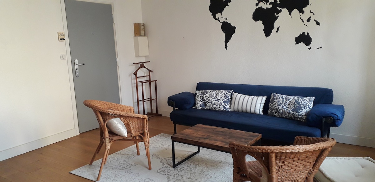 A cozy living area features a blue sofa adorned with patterned cushions and a wooden coffee table. Two wicker chairs are positioned around a light area rug, with a world map displayed on the wall. The space is bright due to natural light from the window.