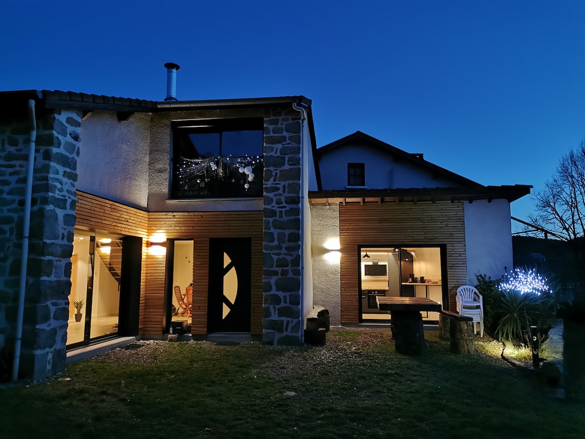 The exterior of the renovated gîte is viewed at dusk, featuring a blend of stone and wooden elements. Soft lighting from the windows illuminates the space. The yard includes seating options and decorative lights, creating a peaceful ambiance against the twilight backdrop.