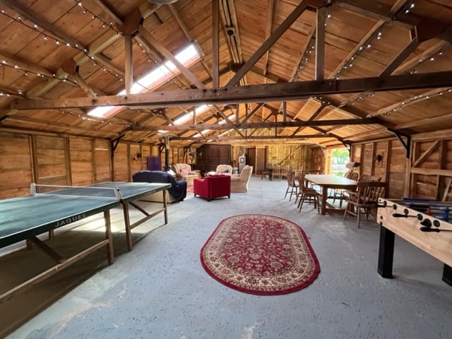 An expansive barn interior features wooden beams and rustic charm, with a table tennis table and a foosball table positioned for recreational fun. Cozy seating areas with sofas are complemented by a dining table surrounded by chairs. Soft lighting enhances the inviting atmosphere.