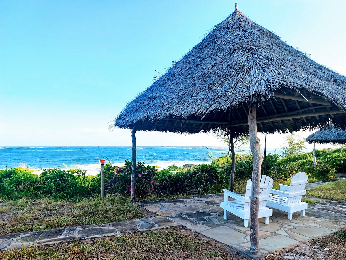 A shaded seating area features two white lounge chairs beneath a traditional thatched roof. The space overlooks a calm ocean, framed by lush greenery and vibrant flowering plants. A stone pathway leads from the chairs to the scenic beachfront.