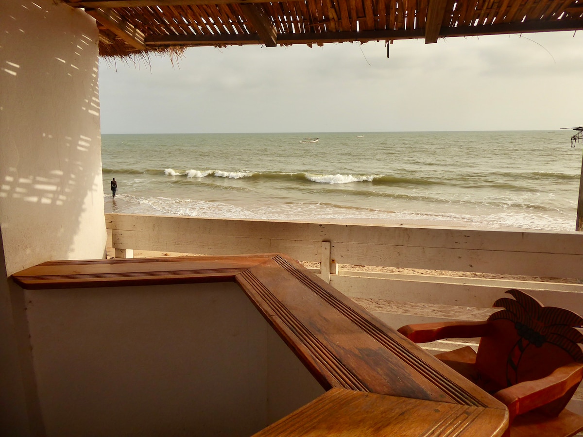 A cozy terrace bar is visible, offering views of the ocean waves. The natural wood bar features a smooth surface, and a carved wooden chair stands nearby. The scene captures the ocean’s horizon under a cloudy sky, enhancing the beachside atmosphere.